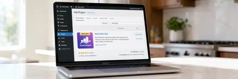 Laptop on a bright white kitchen island displaying the WordPress add new plugin page with the Rank Math SEO plugin card showing a 4.9 star rating as the best SEO plugin for WordPress